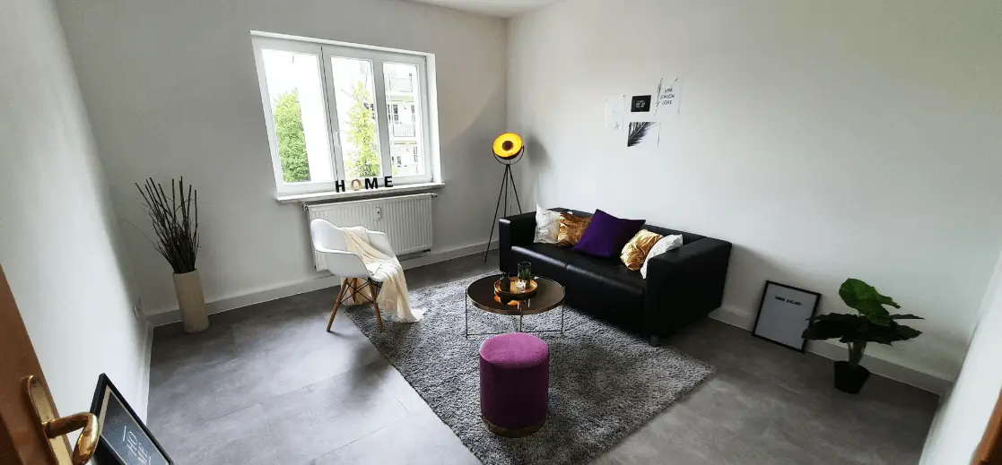 Modern eingerichtetes Wohnzimmer mit dunklem Sofa, violettem Hocker und hellen Wänden. Beispiel für eine professionelle Immobilienpräsentation in Leipzig durch Makler Immo Aslani.